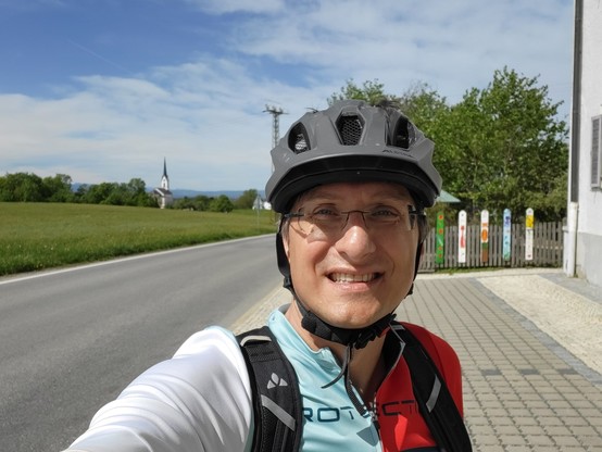 A man is shown wearing a bicycle helmet while standing on a road. The man is smiling. The background consists of a cloudy sky, trees, grass, and a grey road. The man is the main focus of the image, with a bicycle helmet visible on his head. The image suggests that the man is outdoors, possibly riding a bike or preparing to do so. The man is also holding something, likely a phone, as he seems to be taking a selfie. The image captures a moment of joy and outdoor activity.