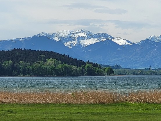 A serene lake is depicted in the image, with a sailboat gently floating in the background. The scene captures the beauty of nature, with lush green grass surrounding the peaceful waters. In the distance, a snowy mountain range can be seen, adorned with trees and fluffy clouds in the sky. The landscape is tranquil and picturesque, evoking a sense of calm and tranquility. The dominant colors in the image are white and green. This outdoor setting showcases a harmonious blend of elements, making it…
