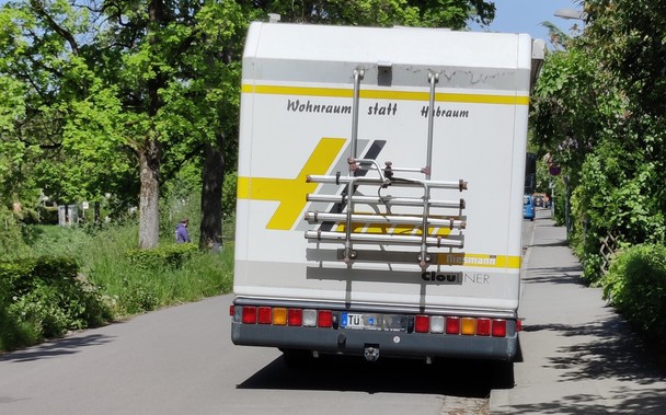 Ein Wohnmobil, das am Straßenrand einer engen Straße parkt und damit knapp die Hälfte der Fahrbahnbreite einnimmt. 
Auf dem Heck des Fahrzeugs ist eine Beschriftung aufgeklebt: "Wohnraum statt Hubraum"