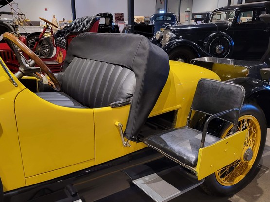 A vintage yellow convertible with a fold-out seat that slides out of the side of the car 