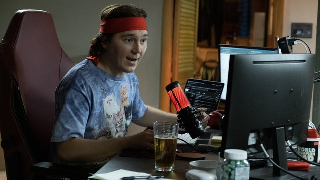 Paul Dano sitzt am Schrebtisch vor drei Monitoren und spricht in ein rot leuchtendes Mikro. Er trägt wein rotes Stirnband und Batikshirt mit Katzen drauf.