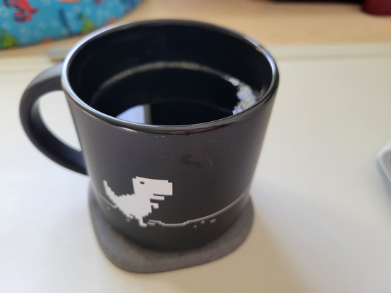Black mug of cold brew coffee on a gray diatomaceous coaster on an off-white desk mat on a maple desk. The mug shows a pixelated dino in white on a black background. The dino is the protagonist T-Rex from The Dinosaur Game, a browser game developed by Google and built into the Google Chrome web browser.