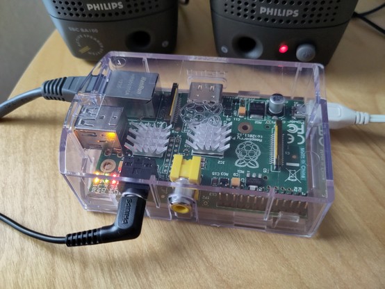 Foto eines Raspberry Pi Model B in einem transparenten Gehäuse. Er ist mit einem Netzwerk-, Strom- und Audiokabel verbunden. Im Hintergrund sieht man ein kleines Paar Lautsprecherboxen stehen.