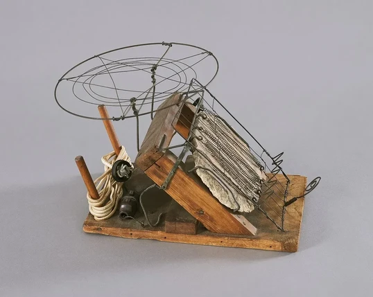 A very rough contraption made of a wooden frame, a stone backpiece, wire supports, and curled wires attached to an electrical cord, for heating the bread. There is a thing that looks a bit like a helicopter landing pad above, possibly for warming buns, or possibly as a second heating element. 