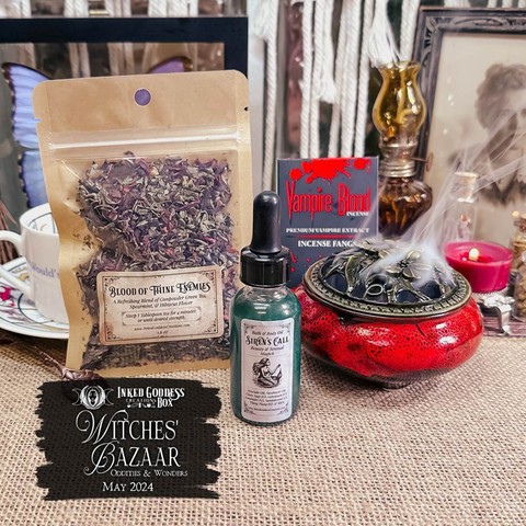 A small pouch of colorful dried herbs is propped up on a teacup. Next to it sits a round, squat red bowl with an antiqued brass lid emitting incense smoke. The final item is a clear bottle with a blue-green liquid inside. These are three of the items in the May 2024 "Witches' Bazaar: Oddities and Wonders" Inked Goddess Creations Box.