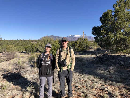 Photo of a smiling woman and man, both wearing binoculars and gray hats with GBBO logos. They are standing in an open pinyon-juniper woodland, with a brilliant blue sky and the snow-covered San Francisco Peaks in the distance.
