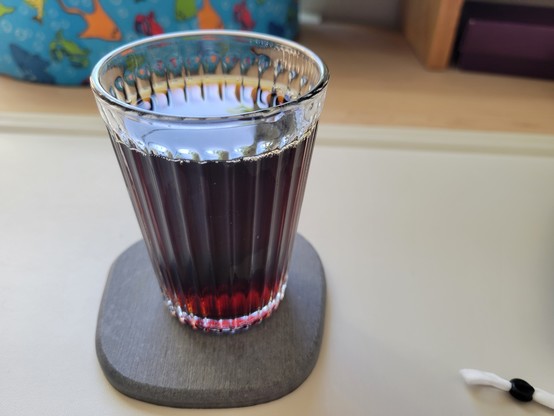Black cold brew coffee in a full, 7-ounce drinking glass (IKEA VARDAGEN - clear with vertical ribs), on a gray diatomaceous coaster on an off-white desk mat on a maple desk.
