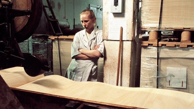 Kati Outinen lehnt mit verschränkten Armen an einer Säule in der Streichholzfabrik. Sie schaut mit leerem Blick vor sich in die Luft.
