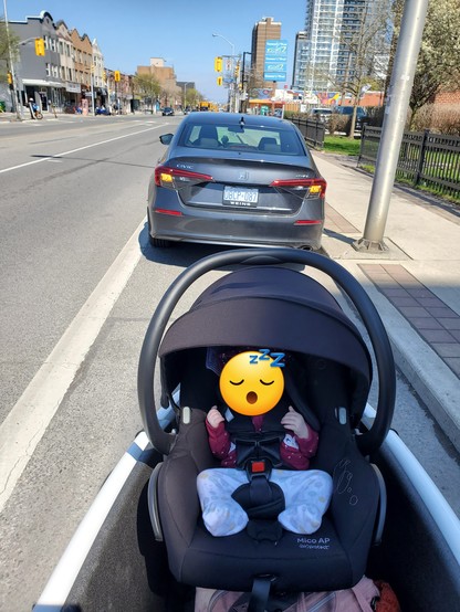 A car stopped in the bike lane on Danforth with 4-way flashers on. Behind the car is a view of the bucket of my cargo bike with a sleeping baby in a car seat.