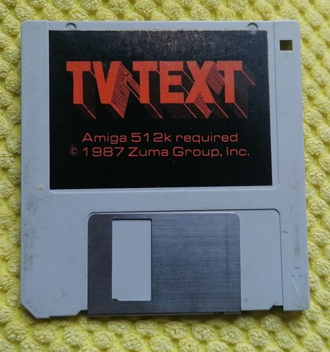 Grey 3.5" floppy disc with black label and lurid, drop shadow orange text that says: TV Text and in smaller orange text: Amiga 512k required copyright 1987 Zuma Group Inc