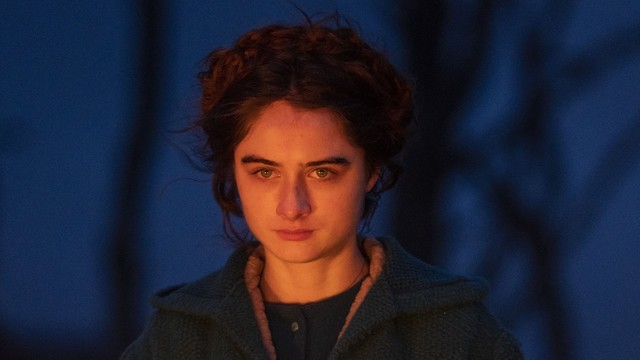 Raffey Cassidy steht im Zwielicht mit einer Flechtfrisur im Wald und schaut desillusioniert drein. Ihr Gesicht wird von einem Feuer angestrahlt.