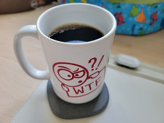 White, mostly full, 15-ounce stoneware mug of cold-brew coffee on a gray diatomaceous-earth coaster on an off-white desk mat on a maple desk. 

The red line-drawing on the mug logo shows initialism "WTF" on a rectangular sign, held by a cartoon character peering over its upper-left corner with one eye wide open and one eye squinting, while emoting a question mark and exclamation point to the right of its head.
