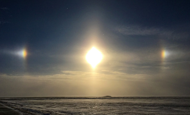 Prismic affect of ice crystals hanging in the air, causing a halo with bright points around the setting sun.  Vista of ice and snow covered wheat fields as far as vision can see. One third of lower ski has clouds. Sun with its dog are slightly above clouds. 

#photography.