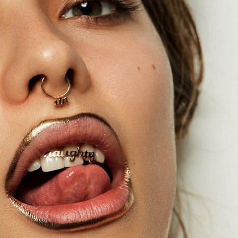 Woman with gold lipliner, gold septum ring, and gold top grillz that read "Naughty" in brush script