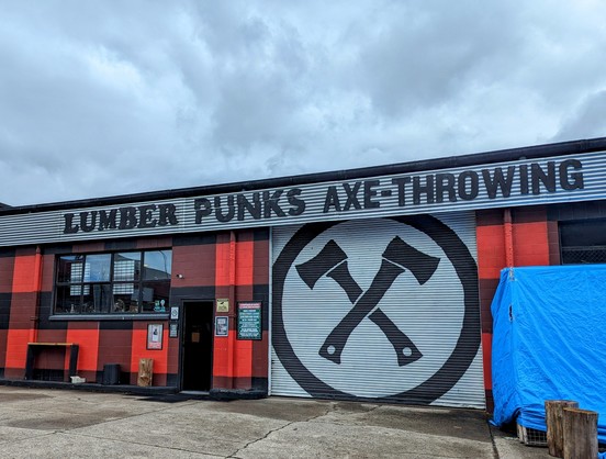 Frontage of an axe throwing venue. 
Cloudy sky above. 
Grey asphalt car park foreground. 
The building is painted in the Macgregor tartan (red & black), has a large "lumber punk axe-throwing" sign running across the top in black letters on roofing tin, and a four metre high white banner with a crossed hatchets design printed in black.
