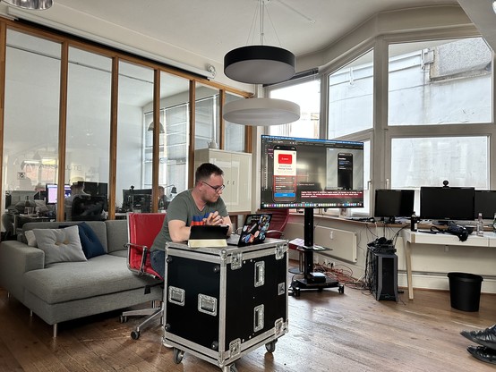 Jan doing his live coding session, sitting on a chair in front of his laptop and iPad with a screen in the background showing the code.