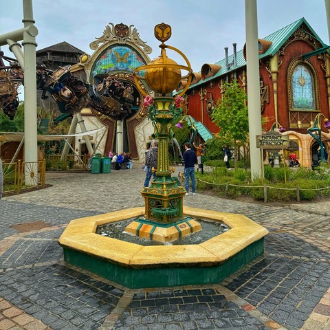 A fountain at the entrance to Ride To Happiness at Plopsaland De Panne, Belgium. On the left, the train travels through a slow first inversion. On the right, the entrance sign can be seen. 
