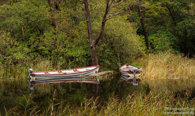 This image features a serene scene of boats floating in a body of water, surrounded by lush green trees in the background. The colors in the image are dominated by shades of brown and black, with a hint of olive green as the accent color. The photo showcases a peaceful outdoor setting, with grass and watercraft visible. The composition includes a variety of boats, including a rowboat with a motor. The overall landscape exudes a sense of tranquility and natural beauty, with a hint of adventure a…
