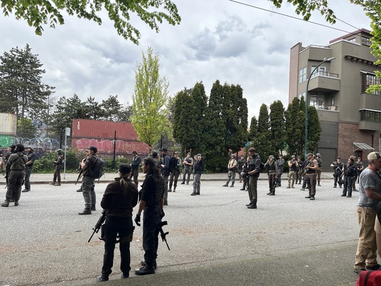 A bunch more movie soldiers standing around with rifles on a street in Vancouver with flowering trees behind them in the sunshine. 