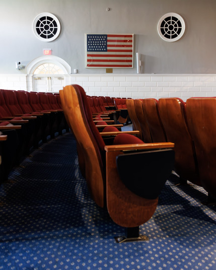 Color photo of several rows of lecture hall chairs. On the opposite wall, are two mullioned round windows, between which is a 46 star American flag in a frame.