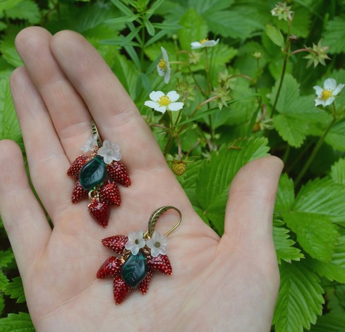 Strawberry earrings displayed on the palm of a hand against a background of wild strawberry plants (vivid green with white flowers). The earrings are an inch long, a cluster of 5 tiny red wild strawberry charms made of enamel. They hang from a bronxe earring hook alonh with 2 small shite flowers and a green glass leaf charm.