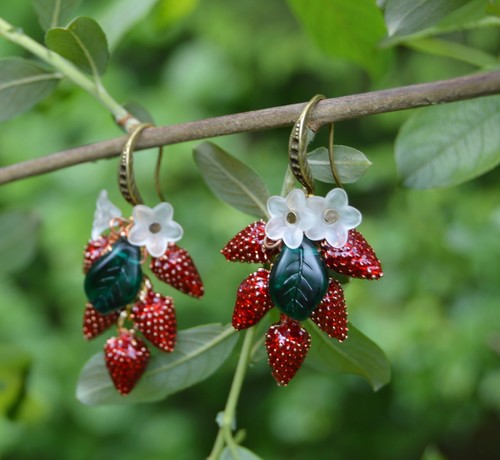 Wild strawberry earrings hanging from a branch, against a blurry background of green leaves. The earrings have 5 small red enamel strawberry charms hanging from a bronze earring hook, along with 2 small white flowers and a green glass leaf.