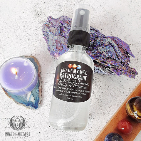A small clear glass spray bottle of "Out of My Way, Retrograde Mist" is propped on a piece of aura kyanite on a white stone surface. The label on the bottle includes the name of the product, its metaphysical properties, and its ingredients. This is an exclusive product from Inked Goddess Creations.