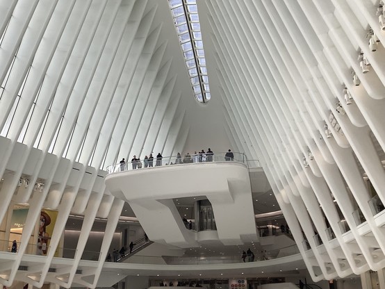 Many people stand on top of a balcony inside a large atrium of white structural members with windows between them that look something like a very angular rib cage.