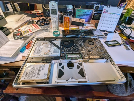 Photo of upside down and exposed bottom half of a 15" MacBook Pro mid 2010. Where the black battery is supposed to go is an empty aluminum area and the bottom half of the touchpad mechanism. Glorious circuitry beams upwards including black dual fans, Intel Core Duo processor, original 500GB hard drive. The circuit board is a wonder to behold. The about-to-be-replaced 2GB RAM chips are embedded next to the battery compartment. 