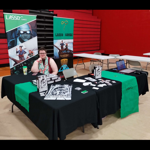 a image of Lasso's final boss con set up! pretty similar to the other posts. Two banners in the back featuring the logo and games. Random quest displayed in the front with a laptop with our games. Random quest boxes, magnets, and mats are also on display. 