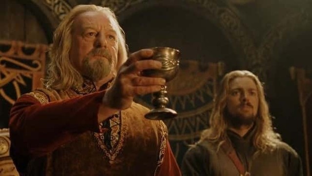 Screenshot from The Lord of the Rings, of Bernard Hill, in his portrayal of King Théoden, as he raises a goblet in toast to those who fell in battle. Behind him is Éomer, his sister’s son, played by Karl Urban, in the Golden Hall of Meduseld. 
