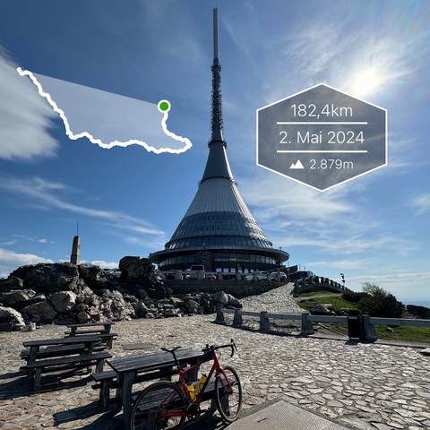 A large tower with a spire atop a mountain, a bike parked beside a bench, rocks, and a clear sky. There's an overlay indicating distance, date, and altitude.