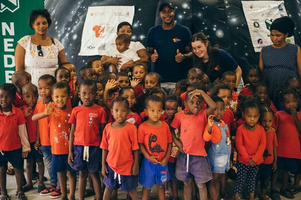 Group of children in red tops with adults standing in the background inside a room, some children are waving, and banners are hung behind on an inflatable planetarium.
