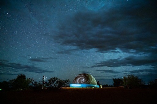 A starry night sky over a landscape with an illuminated blueish dome and a person standing to the side, possibly observing the sky