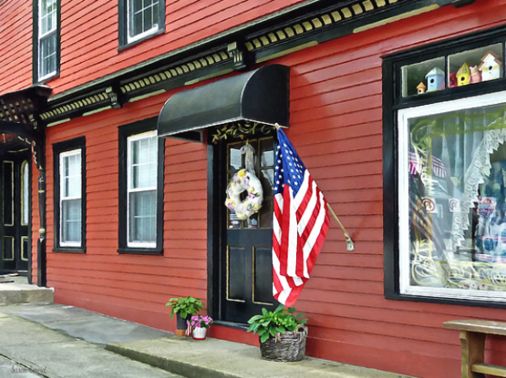 
Cold Spring is a quaint Putnam County village with many antique shops, stores and restaurants. In this image, a vibrant American flag is prominently displayed in front of a red storefront with a black awning. The entrance has a decorative wreath on the door, and there are potted plants adding a touch of greenery at the base of the store's façade.