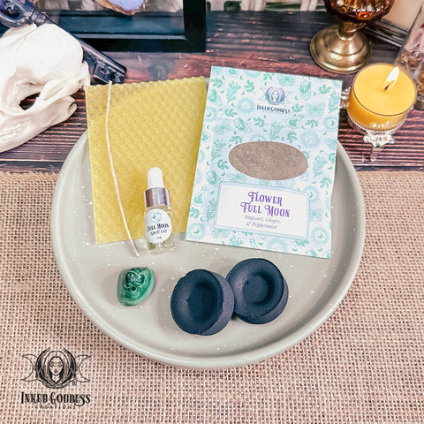 The contents of our exclusive "Flower Full Moon Ritual Kit for the May 23rd Full Moon in Sagittarius" product are arranged on a small tray. They include a sheet of yellow beeswax and a wick; a pouch of Flower Full Moon herbs, including mugwort, ginger, and peppermint; a dropper vial of our exclusive Full Moon Spell Oil; a tumbled malachite gemstone; and two charcoal discs. Presented by Inked Goddess Creations.