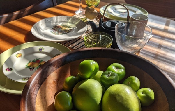 A wooden table striped with sunlight. On it are mixed vintage plates that are pale green or have various designs, and there is a bowl of green apples and tomatillos that I better use before they get smooshy. Probably for chili verde 