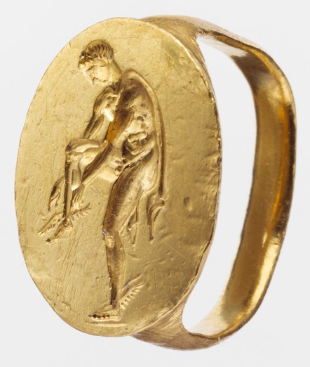 Gold finger ring engraved with an image of Hermes. The broad oval bezel of this heavy gold ring is engraved with an intaglio showing the youthful messenger-god Hermes balancing on his left leg as he fastens a wing to his raised right foot. The god wears a short mantle that encircles his neck and hangs down his back. Winged sandals or boots are a standard attribute of Hermes, but it is unusual to find the wings attached to the figure's ankles rather than to some form of footwear.