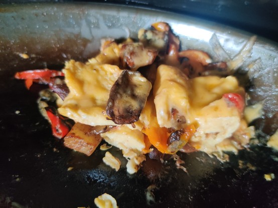A portion of breakfast - cheesy eggs layered with browned potatoes, crisp peppers, and seasoned mushrooms, heaped at the edge of a cast iron pan.