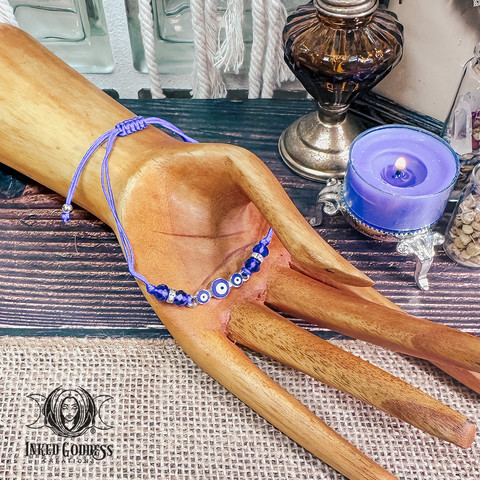 A bracelet rests on an abstract wooden carved hand with its palm up. It features three small evil eye talismans affixed to a silver bezel, silver beads and connectors, blue cord, and blue faceted beads. The bracelet is adjustable via a sliding knot. Presented by Inked Goddess Creations.