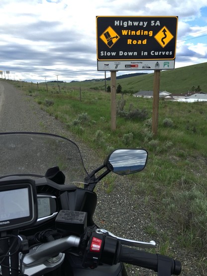 A road sign reading "Highway 5A Winding Road Slow Down in Curves" viewed from a motorcycle handlebar, with grassy terrain in the background.