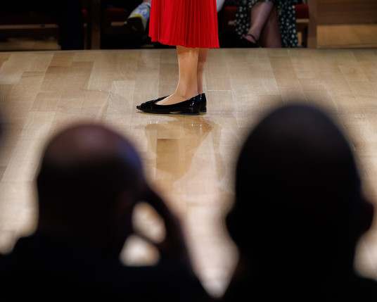 Color photo of the floor of a stage, in the center of which, the side view of the lower part of a woman’s legs are visible from the side. She is wearing a red dress that comes partway down her shins. She is also wearing black patent leather, low-heeled shoes. The stage flooring looks like it is made of maple. In the foreground are the heads of two audience members, out of focus and silhouetted. In the background, the legs of seated audience members are visible.
