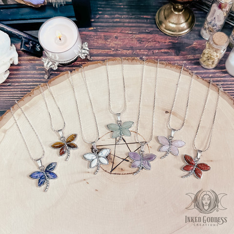 Seven specimens of the "Mixed Gemstone Dragonfly Necklace" product are arranged on a wood slice. The silver dragonfly bezel charms hang from a silver chain, and each dragonfly's four wings are a type of gemstone or mineral, including sodalite, tiger's eye, howlite, green aventurine, amethyst, rose quartz, and red goldstone. Presented by Inked Goddess Creations.