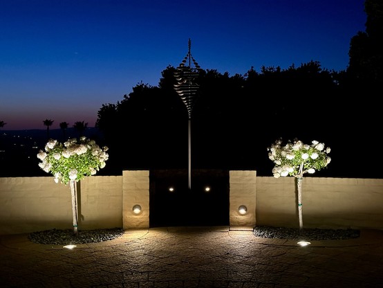 Paved area with two white rose bushes in front of a wall with a gateway to an illuminated wind sculpture.