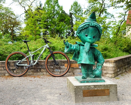Rechts in Bild die Skulptur des Sandmännchens, links dahinter steht ein graues Mountainbike an eine kleine Mauer gelehnt. 