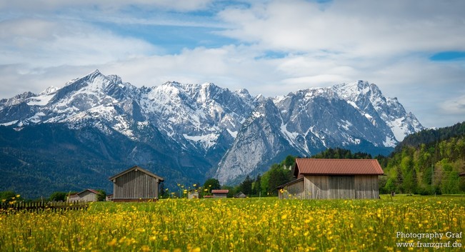 A picturesque scene of a rural landscape featuring a group of buildings surrounded by a vast field. In the background, majestic mountains rise up against the sky, creating a stunning backdrop.  Two prominent objects stand out in the image: a tree in the foreground and a house slightly off-center. The composition evokes a sense of tranquility and natural beauty, with hints of a springtime atmosphere. This serene setting captures the essence of a peaceful countryside retreat, with a touch of alpi…