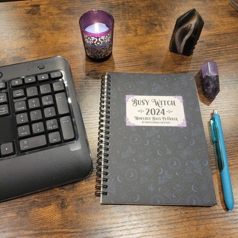 A dark spiral-bound journal sits on a wooden desk top next to a computer keyboard, a pen, two crystals, and a burning candle. The text on the front of the journal reads "Busy Witch 2024 Monthly Date Planner". This is an exclusive product from Inked Goddess Creations.