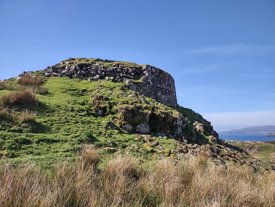 The bottom remains of a dry stonewall roundhouse at the top of a rocky knob of land, with short green grass and mosses and tufts of long brown grass from last year. It probably was originally 50 feet or so tall, but only the bottom 5 to 10 feet remain.