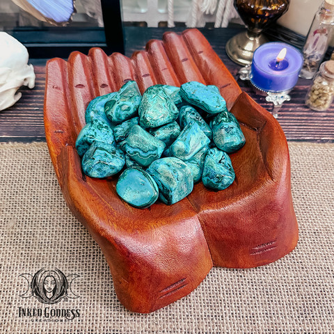 A variety of Malacholla Tumbled Gemstones rest in cupped, carved wooden hands. The specimens are irregular small nuggets, each made of many layers of Malachite and Chrysocolla, resulting in free-flowing blue and green lines and planes. Presented by Inked Goddess Creations.