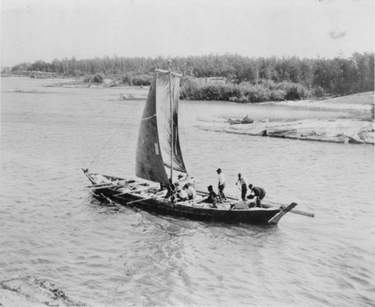 A York Boat under sail along a rock-bound river bank, the steersman at the rear of the boat bending to peer under the sails to see where he is going. 